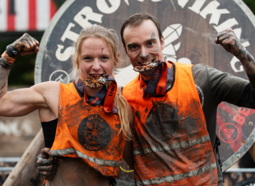 een man en een vrouw die de utlra viking hebben overwonnen, ze laten hun spierballen zien en bijten op hun medaille